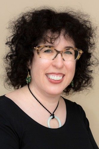 Jennifer Mass smiles at camera for picure wears a pair of tortoise shell eyeglasses, dark top and black necklace with a cricular ivory pendant