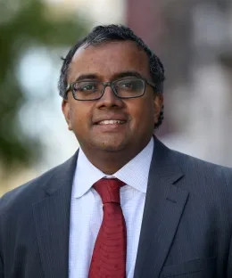 Shyamkrishna Balganesh's Headshot.  He wears a dark rimmed eyeglasses, red tie, white button down colored shirt and a charcoal blazer jacket. 